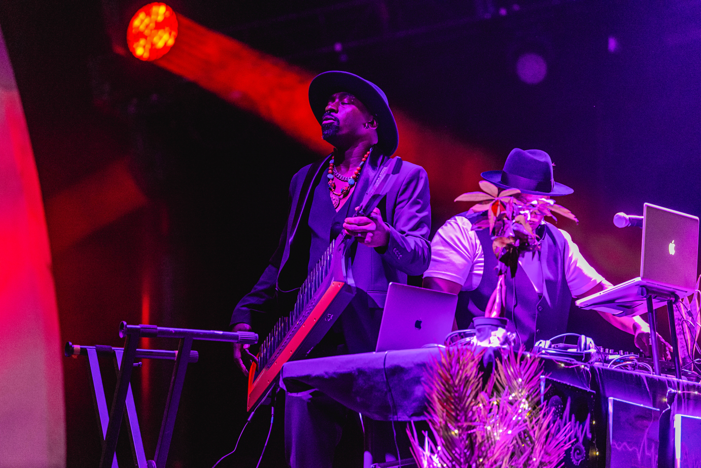 A man in a fedora and suit jacket plays guitar, his eyes closed. To his side, a man in a hat works DJ equipment.